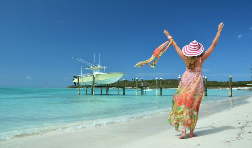 boat-rental | Hideaways Beach Resort Exuma, GeorgeTown Vacation Rentals Woman in a colorful dress and striped hat raises her arms on a white-sand tropical beach with a boat in the distance.