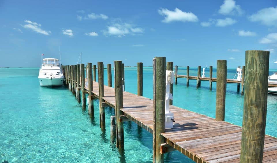 boat-tour | Hideaways Beach Resort Exuma, GeorgeTown Vacation Rentals Long wooden pier extends over clear turquoise water toward a white boat anchored on the left.