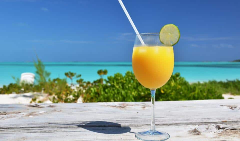 chat-n-chill | Hideaways Beach Resort Exuma, GeorgeTown Vacation Rentals Glass of orange juice with a lime wedge on a weathered wooden table at a sunny beach. The turquoise sea is in the background.