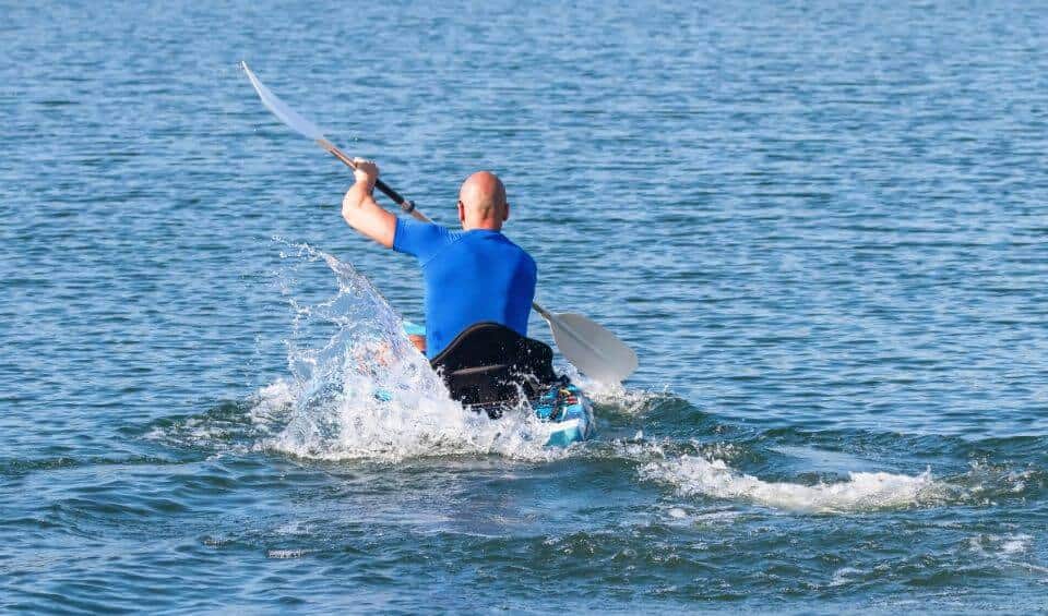 unique-activities | Hideaways Beach Resort Exuma, GeorgeTown Vacation Rentals Back view of a person in a blue shirt paddling a kayak on open water, with the paddle raised mid-stroke over splashing waves.