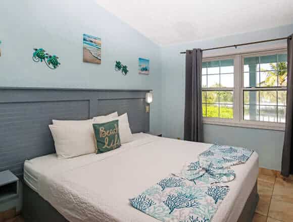 Beach-themed hotel room with a double bed, teal headboard, white linens, and coral-patterned blue throws; window with curtains and palm trees outside.