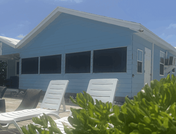 Blue beach house with a row of screened porch windows, white trim, and a white door, seen from a sunny deck with lounge chairs and greenery in the foreground.