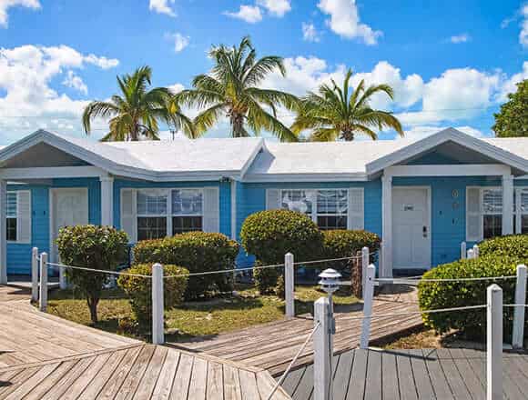 Blue seaside cottages with white trim and palm trees lining a wooden boardwalk under a bright sky, coastal resort vibe.