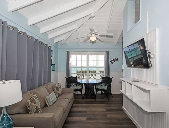 Coastal-style living room with light blue walls, white vaulted ceiling, beige sofa, and seating by a large window.
