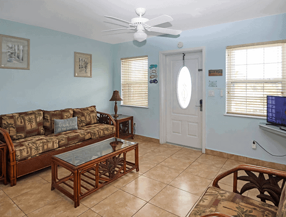 Cozy living room with blue walls, wicker sofa set, glass coffee table, and a front door with an oval window. Ceiling fan above and blinds on the windows.