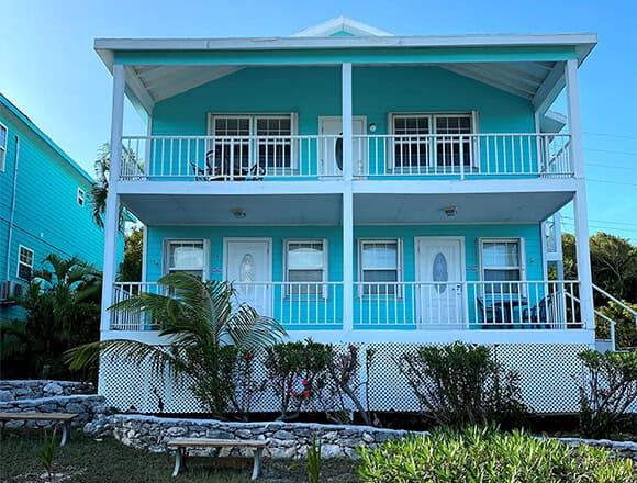 Two-story turquoise house with white railings and balconies, front lawn with palm trees and stone planter.
